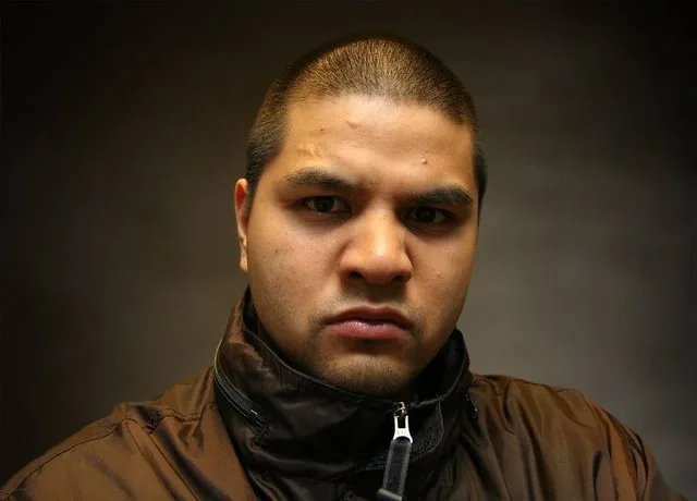 Studio portrait of a young man in a brown zip jacket against a dark backdrop, looking straight down the lens